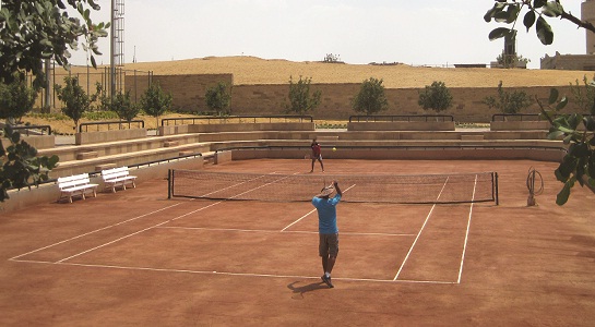 Tennisplatz im Freien mit Sandbelag, ein Spieler beim Aufschlag, leere Tribünen und Wüstenhügel.
