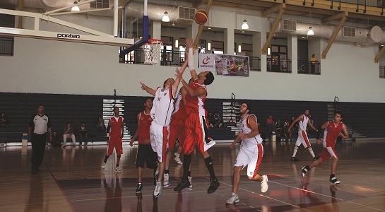 Hallenbasketballspiel: Spieler springen beim Rebound unter dem Korb, Tribünen im Hintergrund.