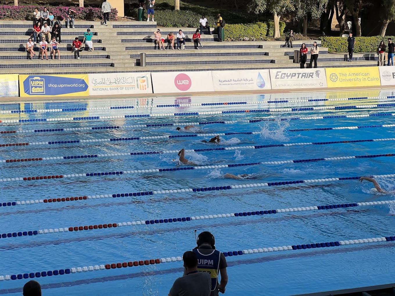 Freibad-Schwimmbecken während eines Rennens, Bahnen mit Leinen markiert, Zuschauer sitzen auf den Stufen.