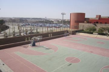 Leeres Mehrzweckfeld im Freien mit Basketballkörben, Blick auf Campusgebäude und Parkflächen.