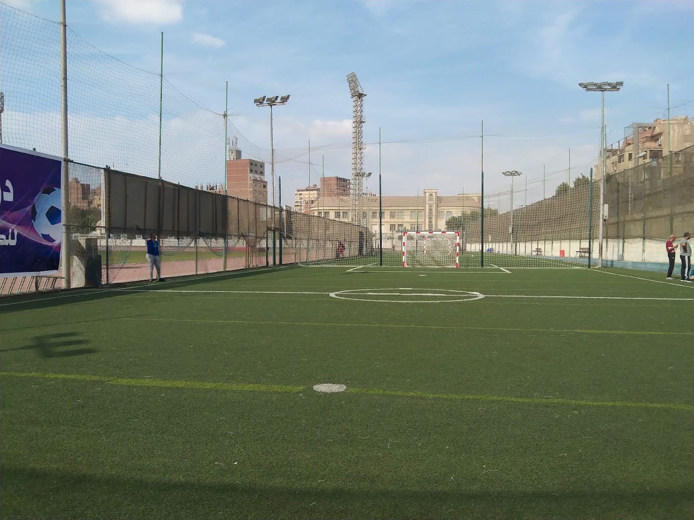 Fenced artificial-turf mini football pitch with a goal and floodlight masts.