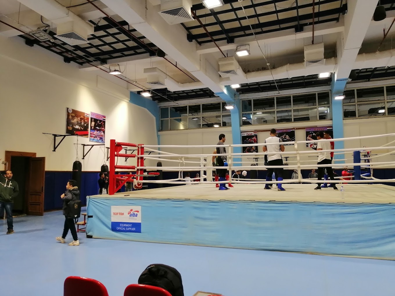 Indoor boxing gym with a full-size ring and athletes training nearby.
