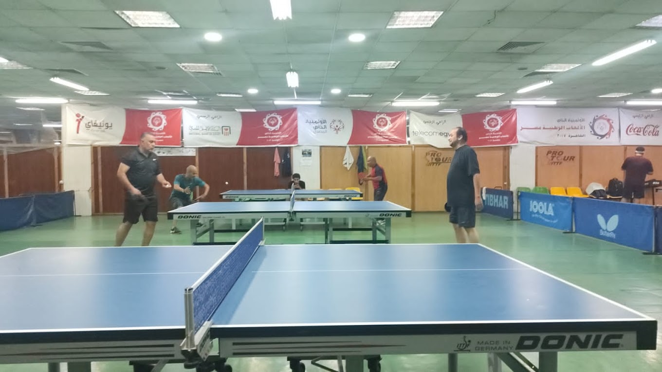 Table tennis hall with multiple tables and players practicing behind barriers.