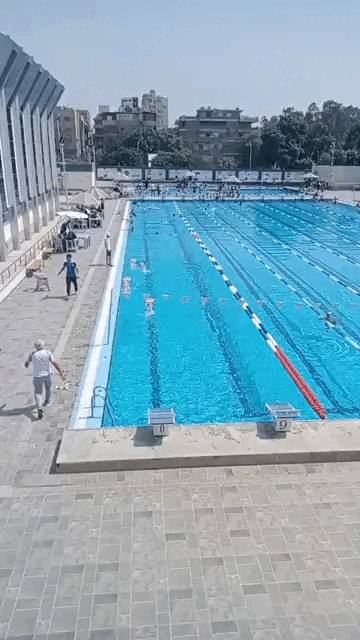 Elevated view of an outdoor lane pool with starting blocks, clear markings, and staff along the pool deck.