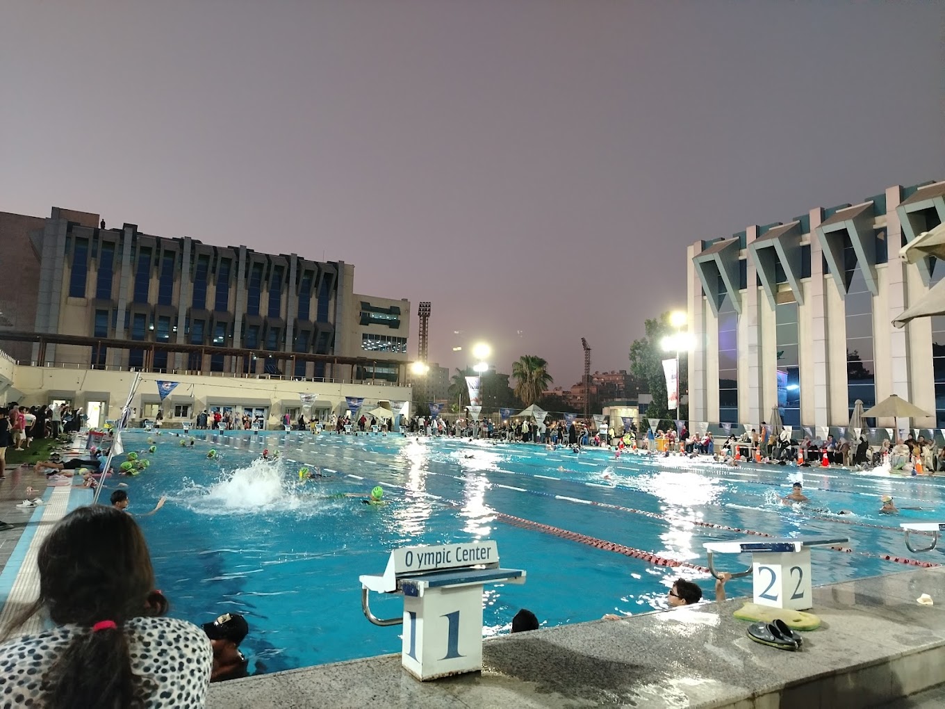 Busy evening session at an outdoor pool under floodlights, swimmers in lanes with people gathered on deck.
