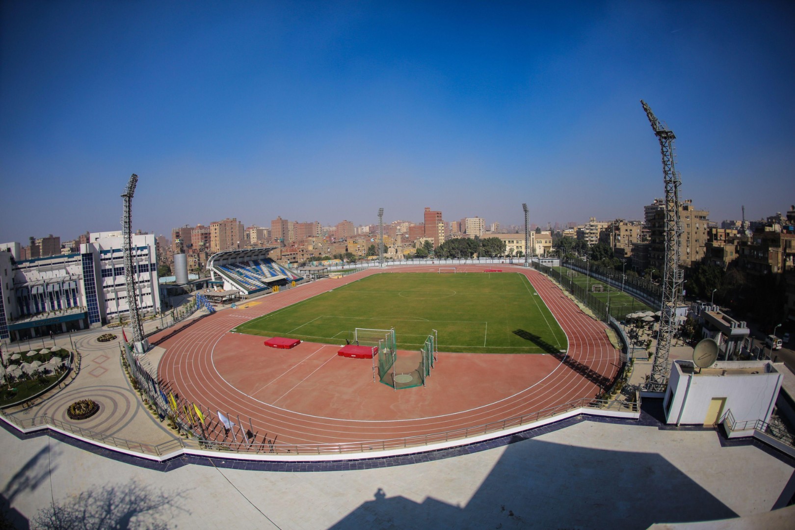 Aerial view of red running track around green field, empty stands and city skyline under blue sky, training camp venue
