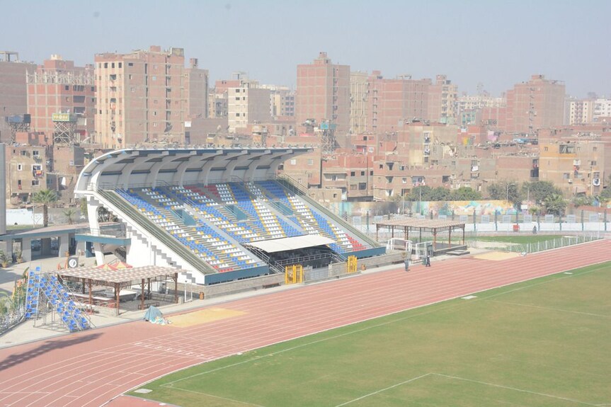 Roofed grandstand with blue and yellow seats beside a red running track and green field; urban backdrop, training camp