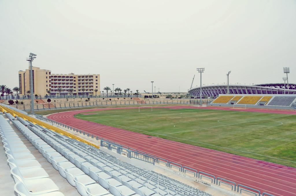 View from white bleachers of red athletics track and green infield, floodlight poles and modern stadium, training camp