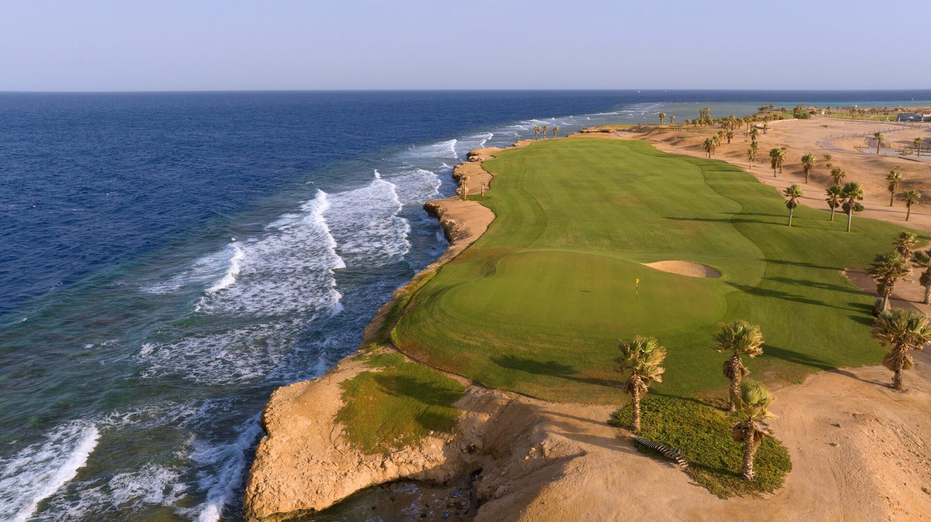 Aerial view of a clifftop green above ocean waves, with flagstick and bunker.
