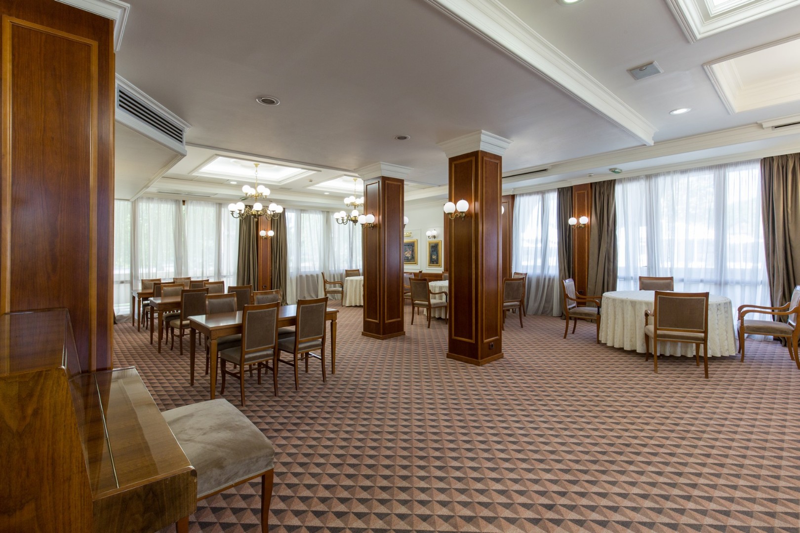 Spacious dining or lounge hall with tables, chairs, large windows and chandeliers.