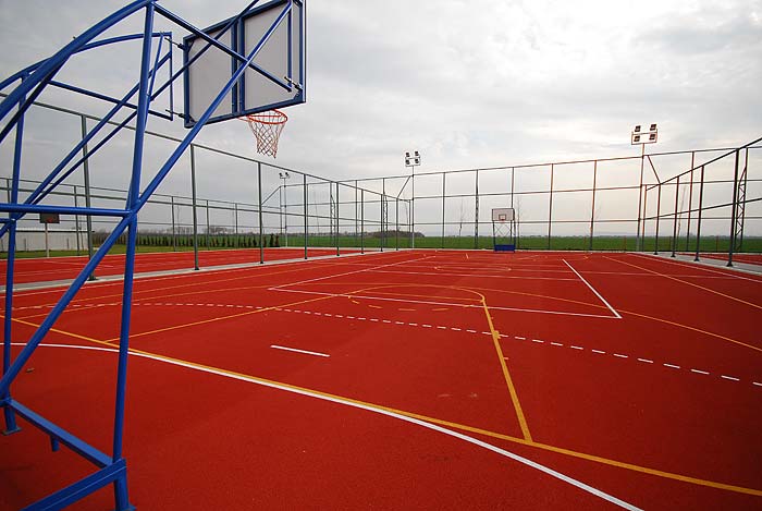 Outdoor multi-sport court with basketball hoops and marked lines on a red surface, surrounded by fencing.