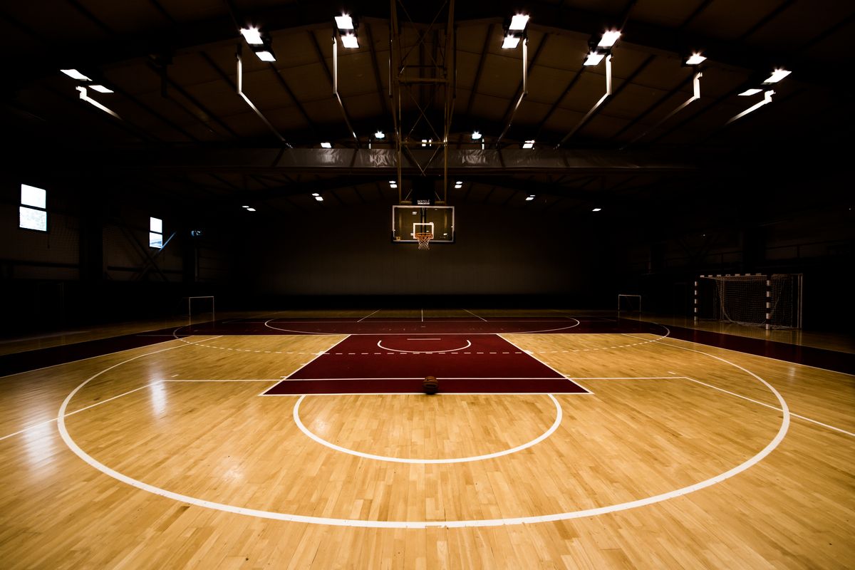 Dimly lit indoor basketball court with hoop and marked hardwood floor.