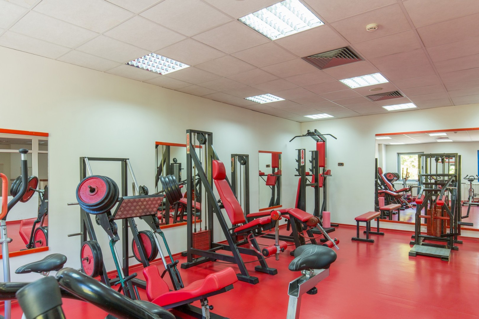 Indoor gym with weight machines, benches and mirrors on red flooring.