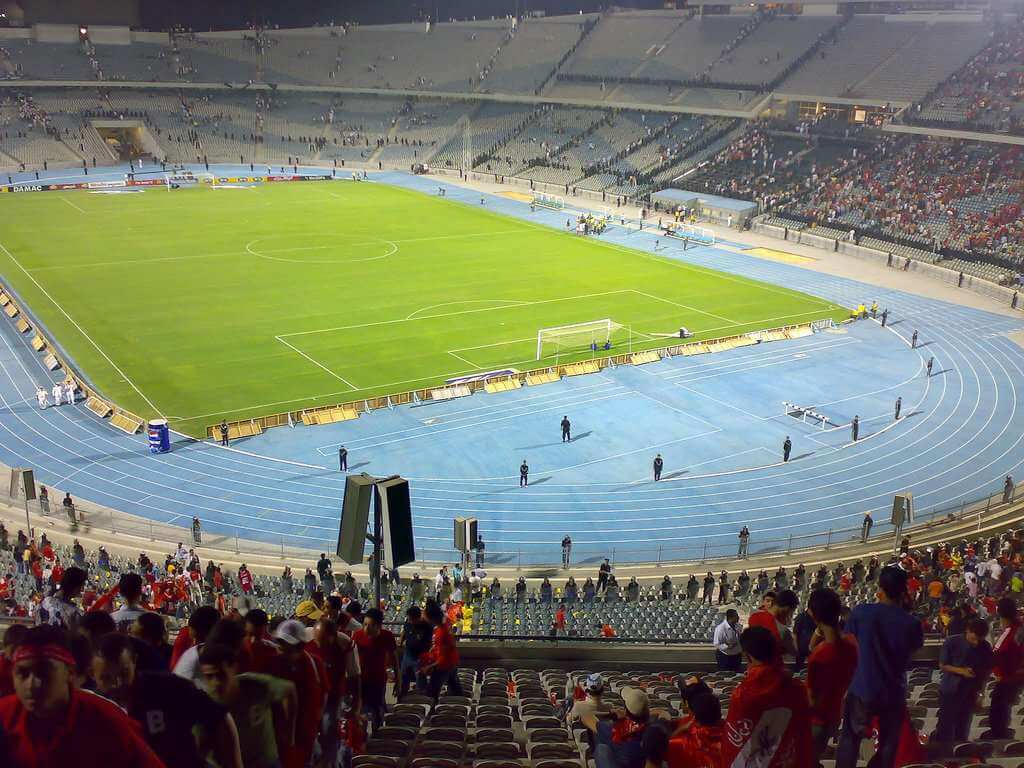 Tribünen mit Zuschauern neben blauer Laufbahn und Fußballfeld im Stadion.