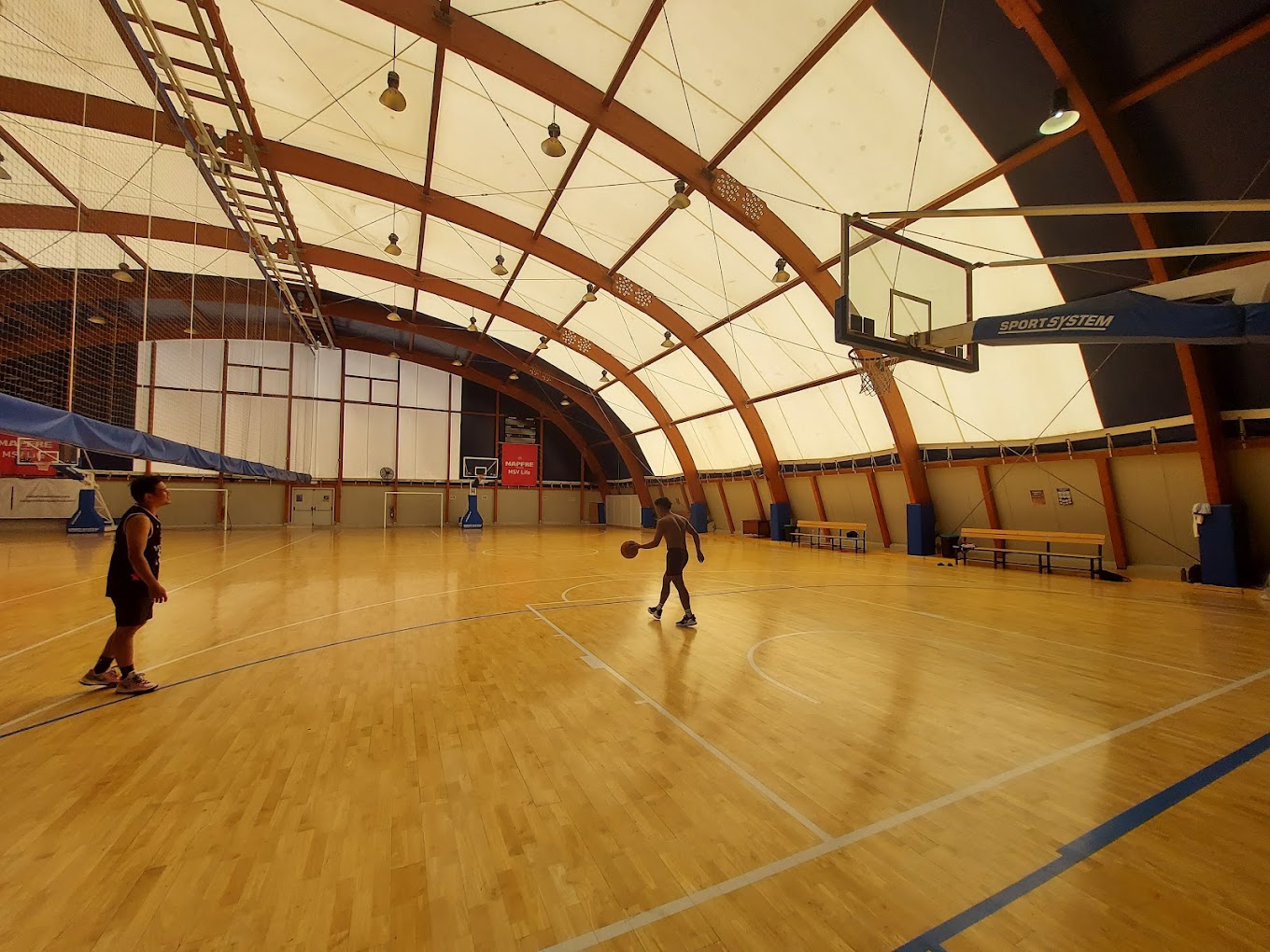 Indoor basketball court with arched roof; two athletes on the court with a ball during practice.