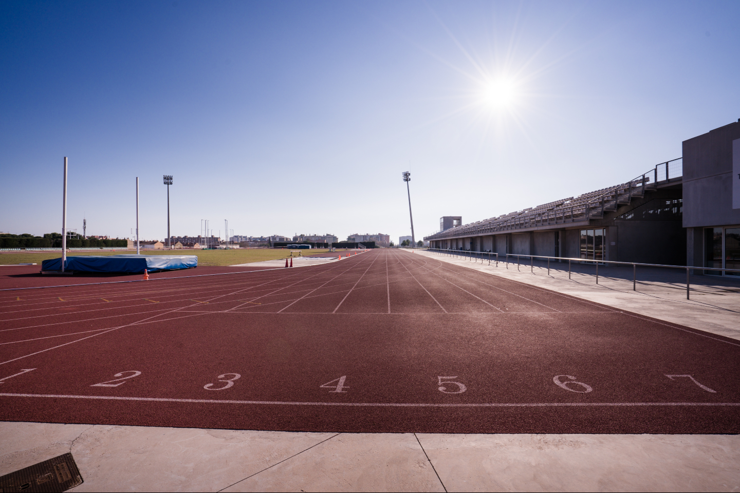 Sonnige Sprintgerade mit Bahnenmarkierungen, Tribüne und einer Hochsprungmatte.