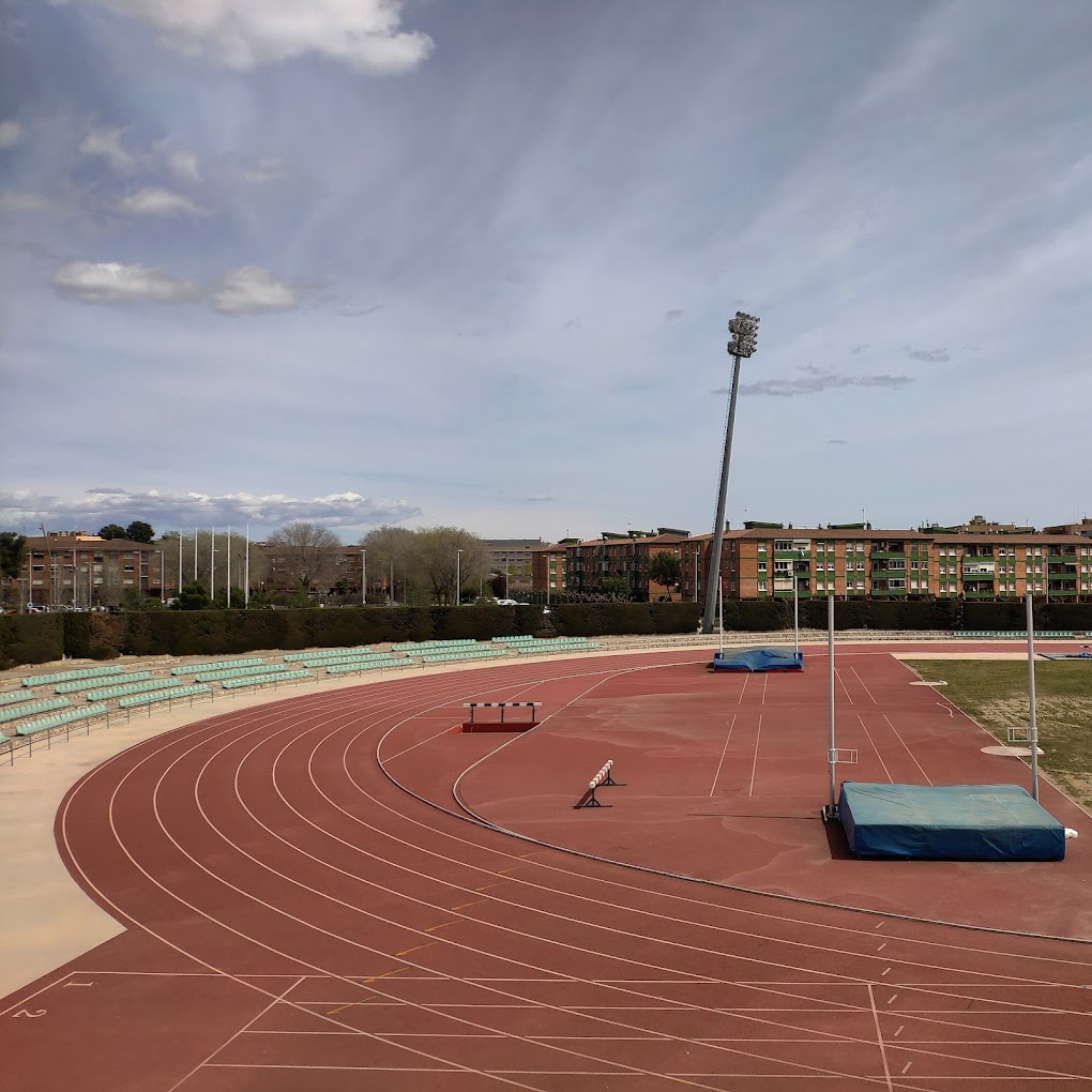 Outdoor-Leichtathletikbahn in der Kurve mit Hürden und Hochsprungmatten neben leeren Rängen.