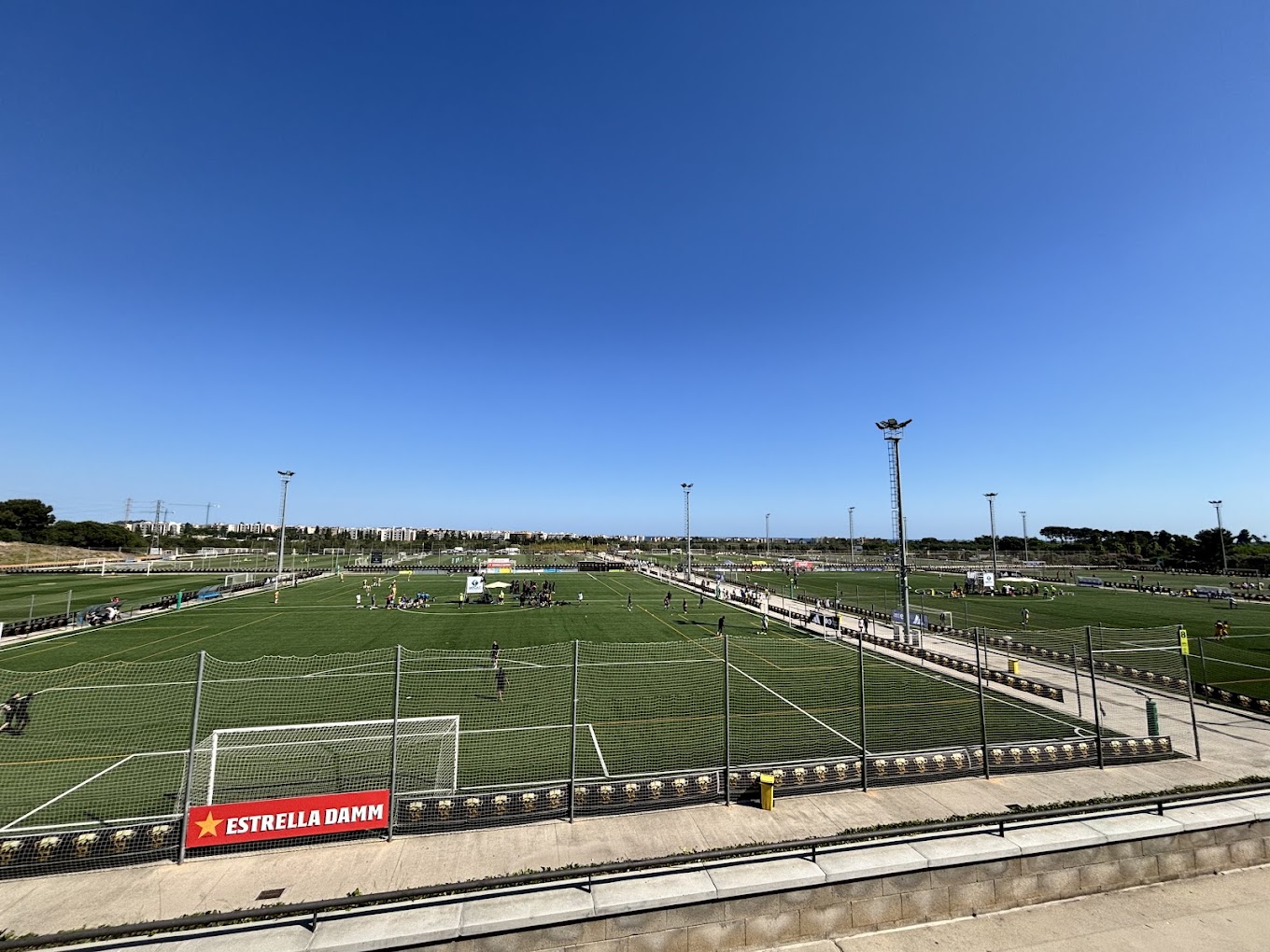 Panorama einer großen Fußballanlage mit mehreren Plätzen und hohen Flutlichtmasten unter klarem Himmel.