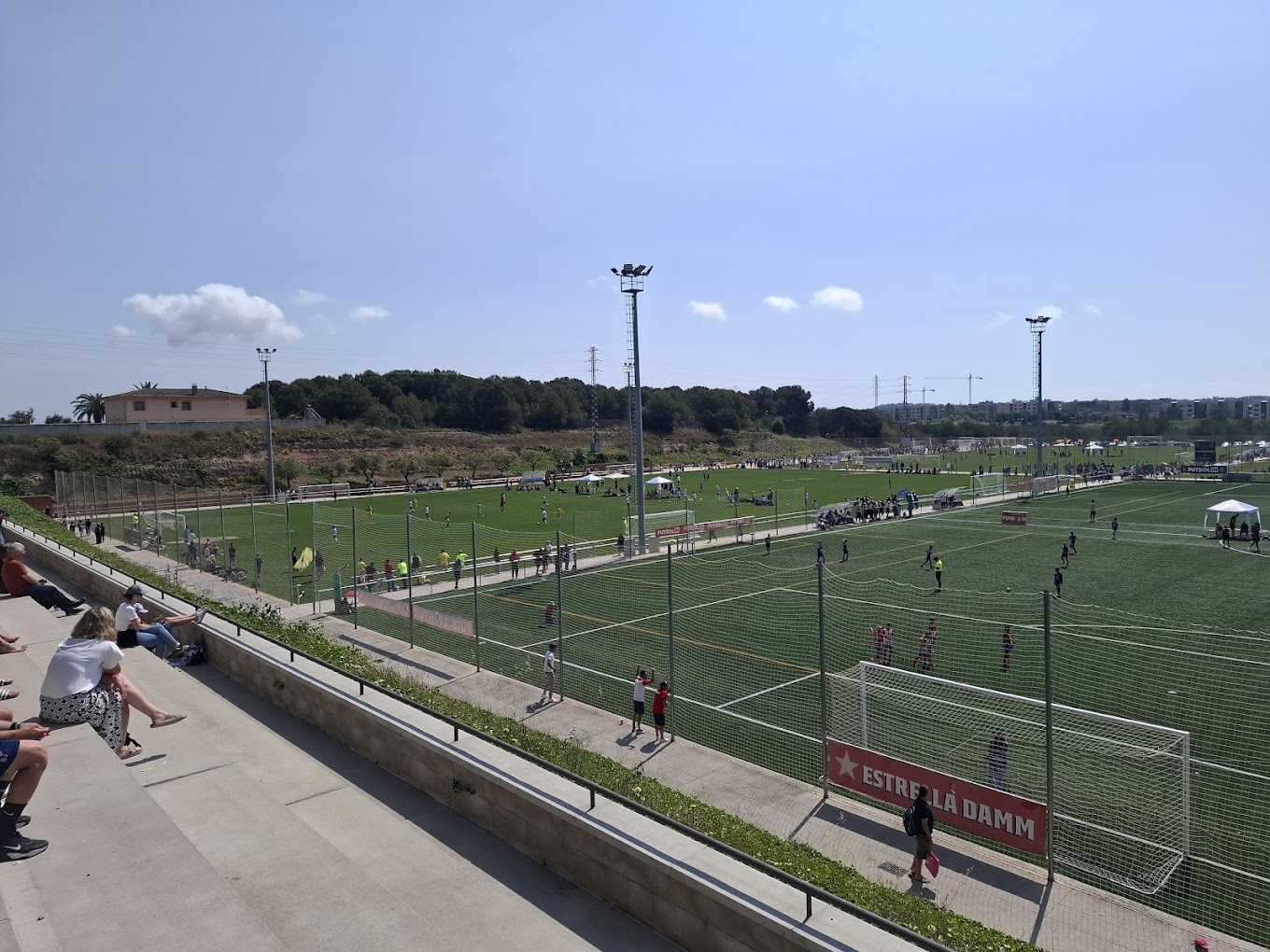 Blick von der Tribüne auf mehrere Fußballplätze mit Flutlicht und Zuschauern entlang der Seitenlinie.