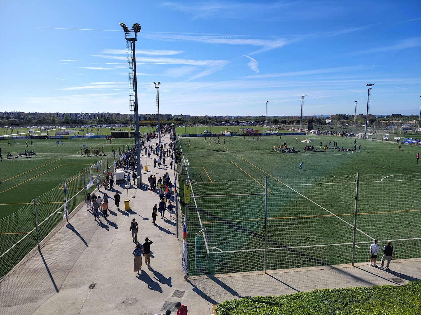 Erhöhte Ansicht eines Fußballkomplexes mit mehreren Plätzen und zentralem Weg mit Besuchern.