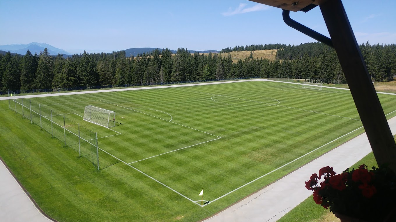 Panoramic view of a full-size grass football pitch beside forested hills.
