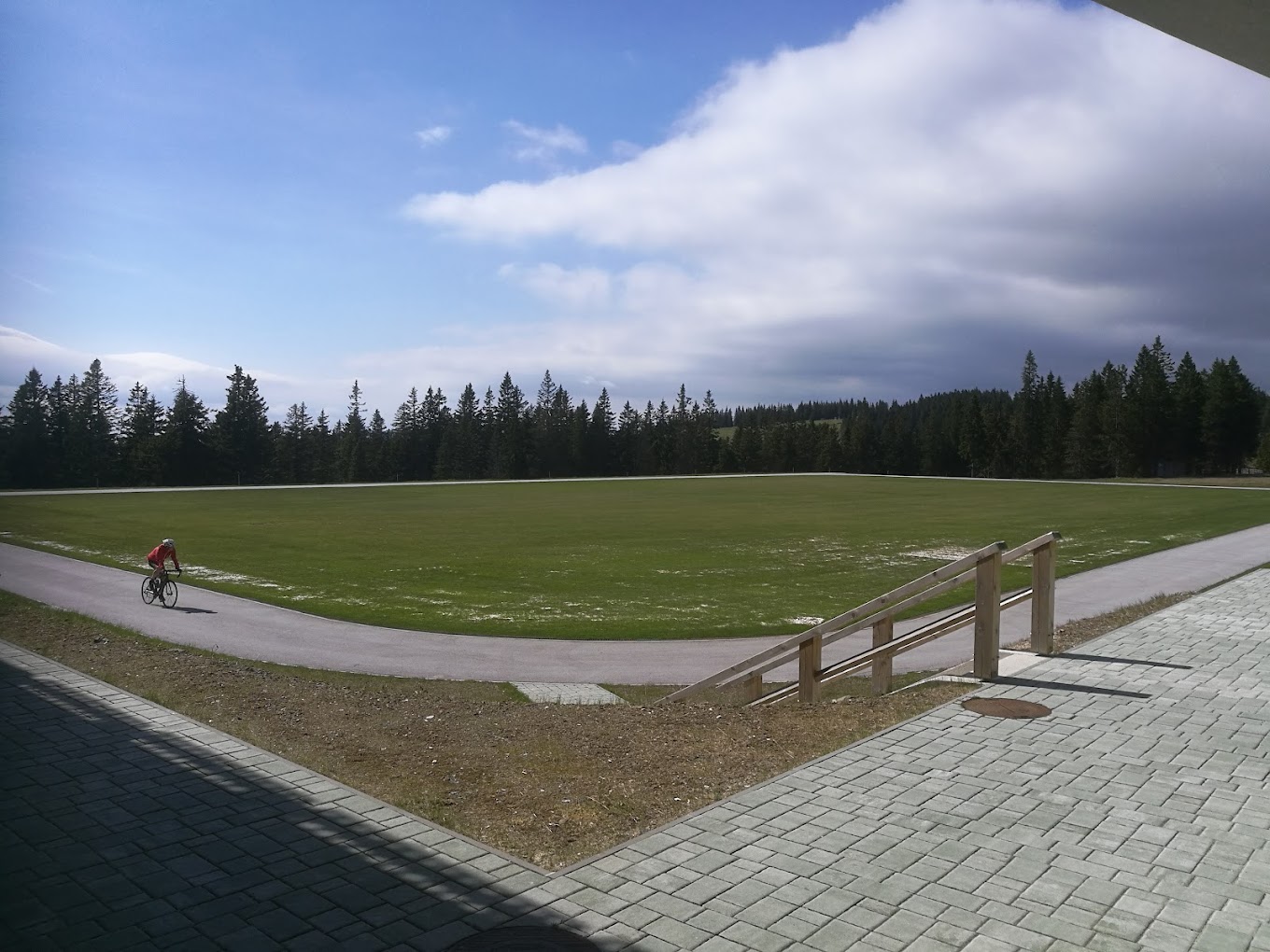 Grass football field encircled by a paved path, with forested hills and a cyclist nearby.