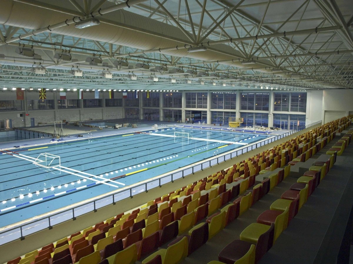 Wide view of an indoor water polo pool with goals and spectator seating.