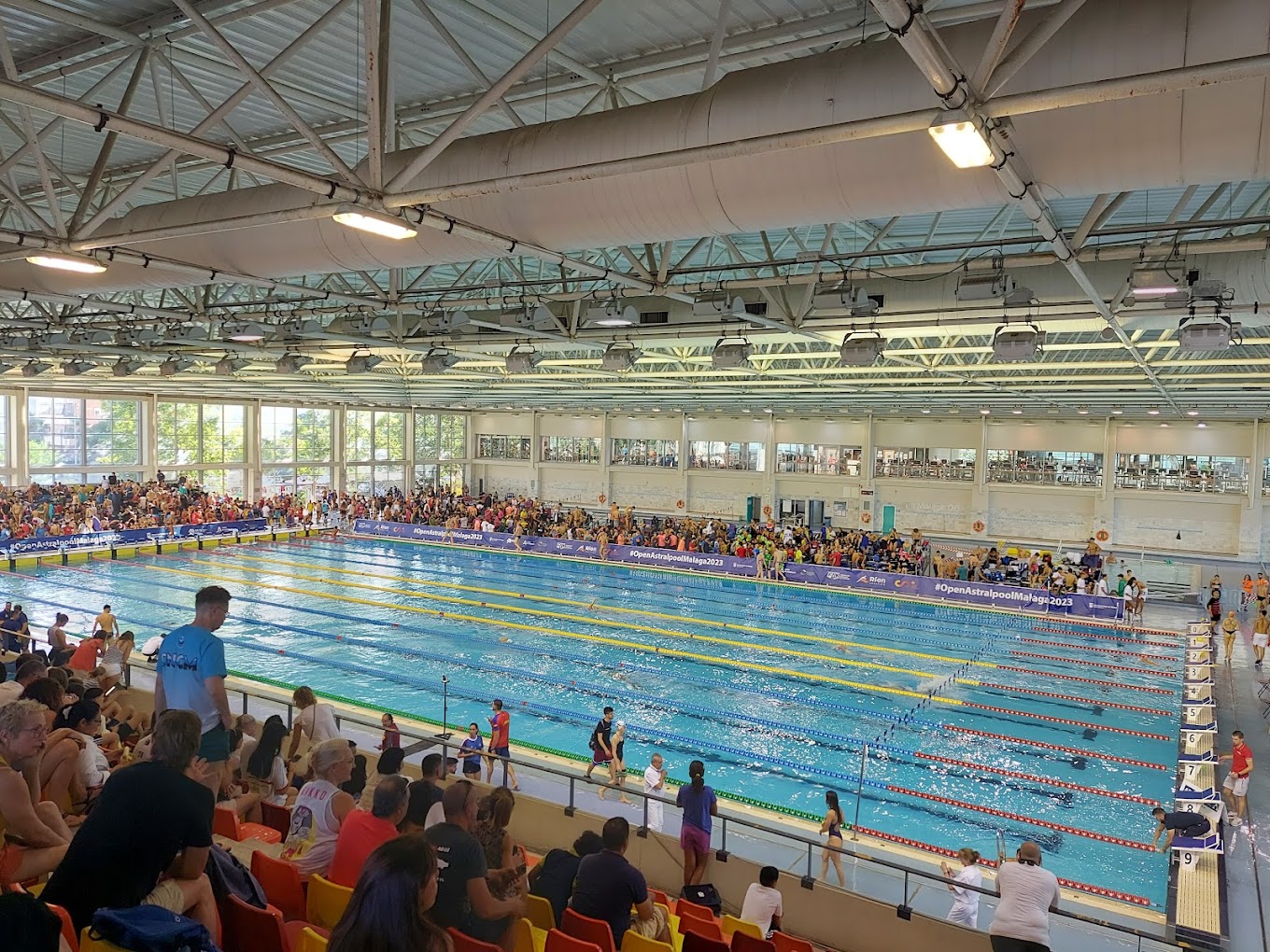 Indoor competition pool during an event, viewed across rows of spectator seats.