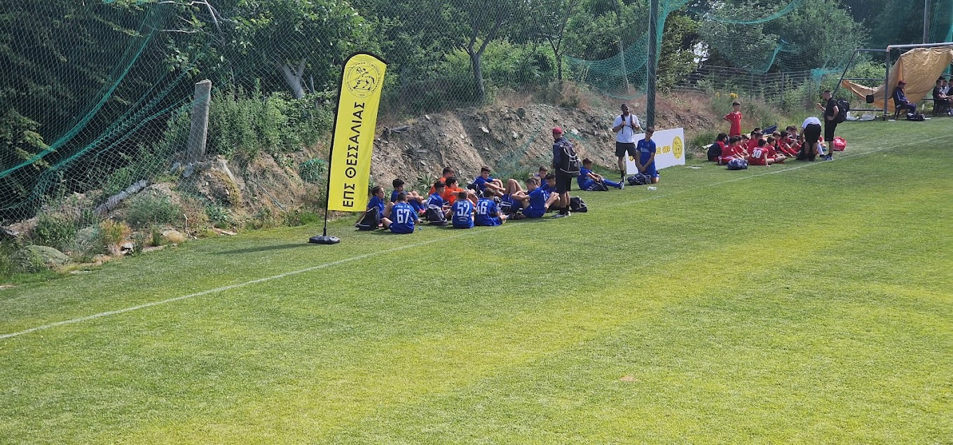 Youth players and coaches seated along the sideline of a football pitch.