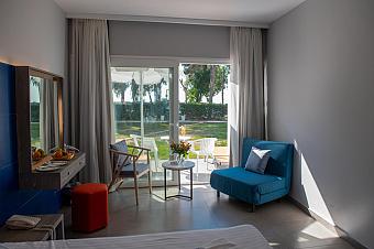 Bright hotel room with blue and gray furniture, a terrace view of a green garden, and a small outdoor seating area