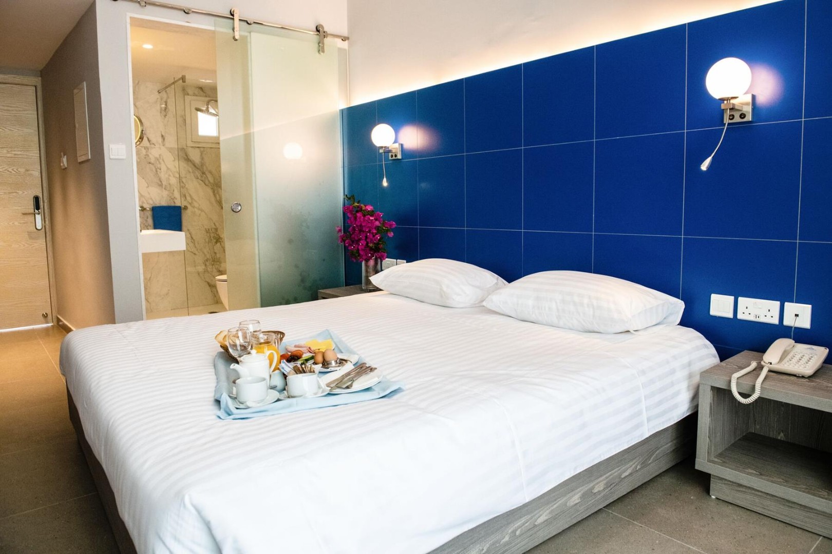 Cozy room with double bed, blue wall, and breakfast tray beside a marble bathroom entrance