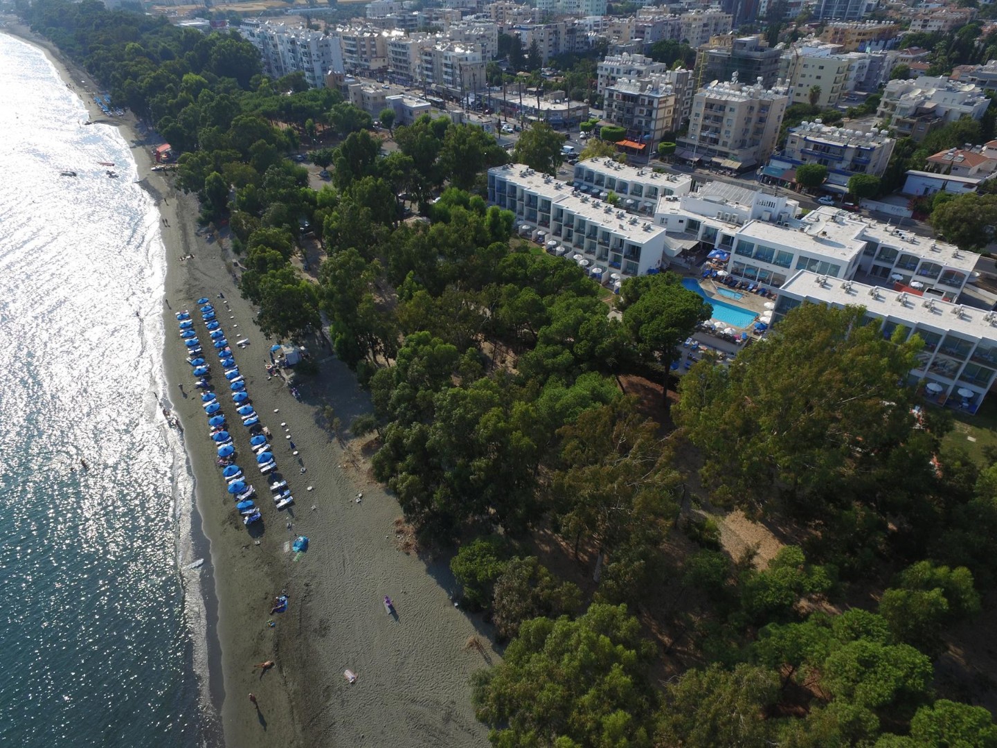 Aerial view of beachfront hotel complex with rows of blue umbrellas and lush trees along the coast
