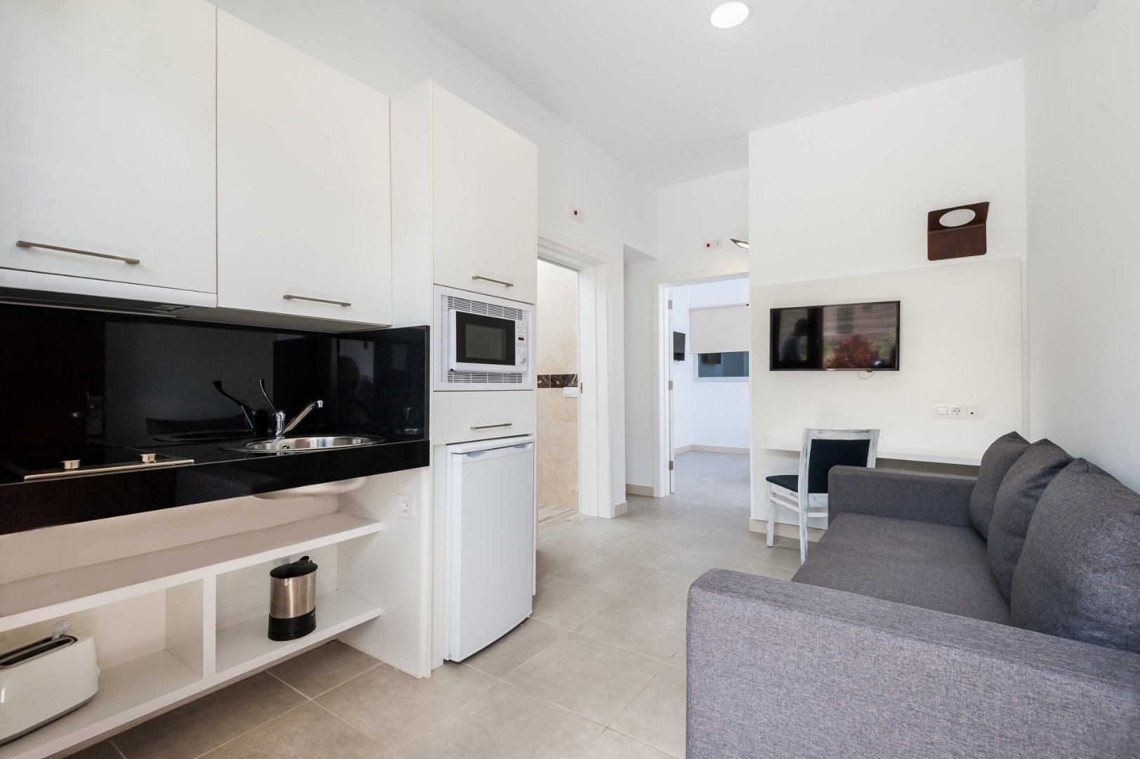 Compact kitchenette with microwave and mini fridge beside a gray sofa and wall-mounted TV.