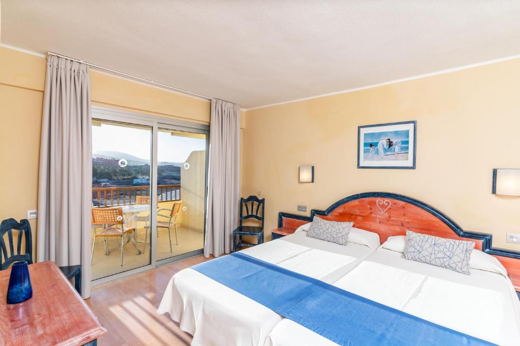 Hotel room with twin beds, wooden desk, and balcony seating visible through sliding doors.