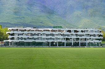 Hotel building facing a green football pitch with mountains behind, serving as accommodation for a sports training camp