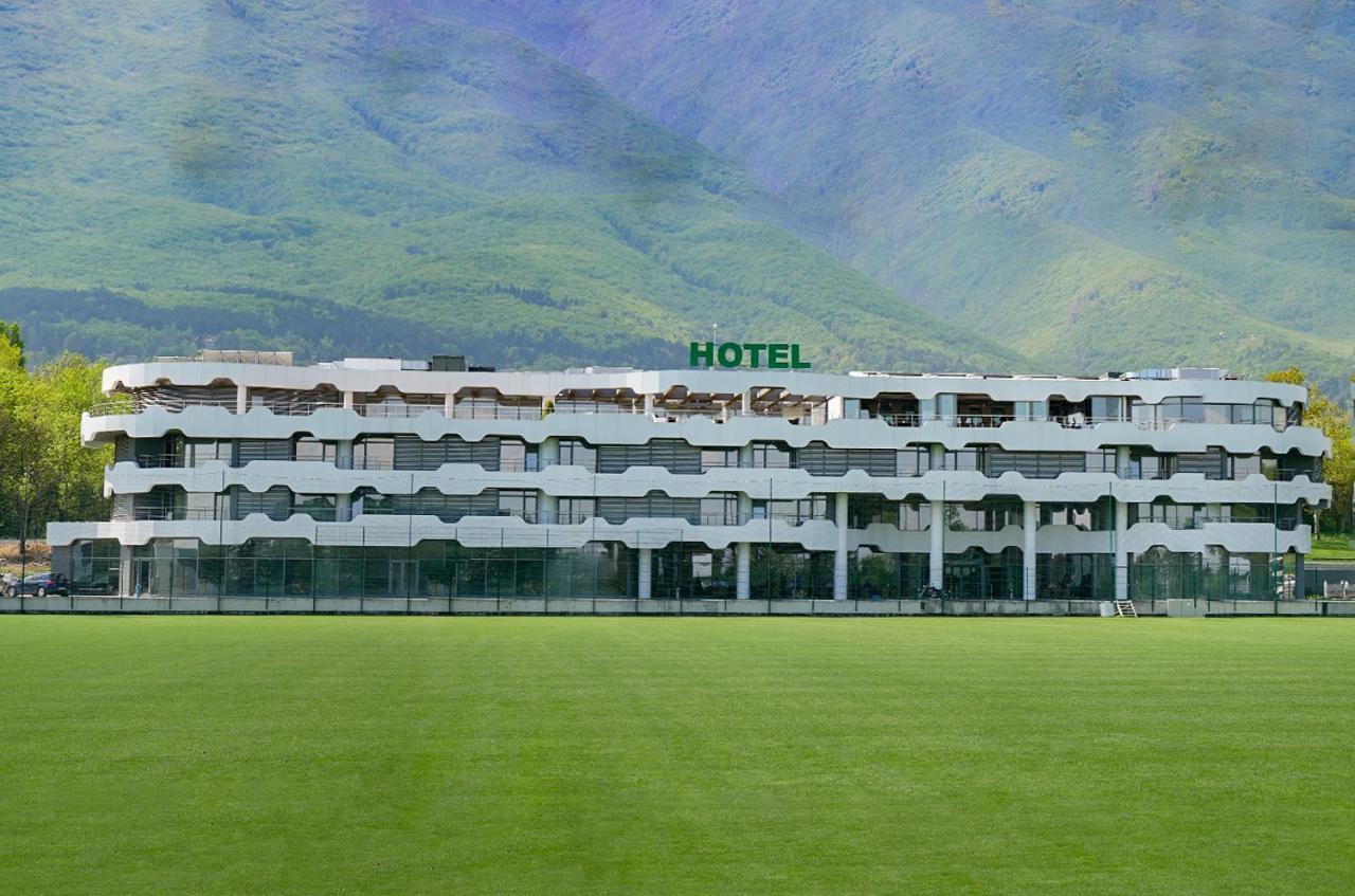 Hotel building facing a green football pitch with mountains behind, serving as accommodation for a sports training camp