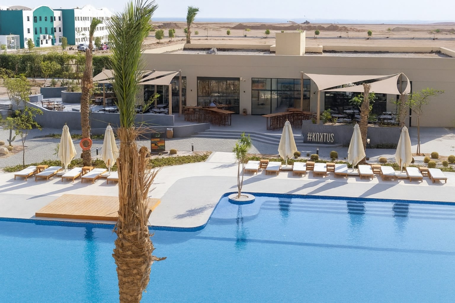 Outdoor pool with sun loungers, umbrellas, and palm trees near resort building