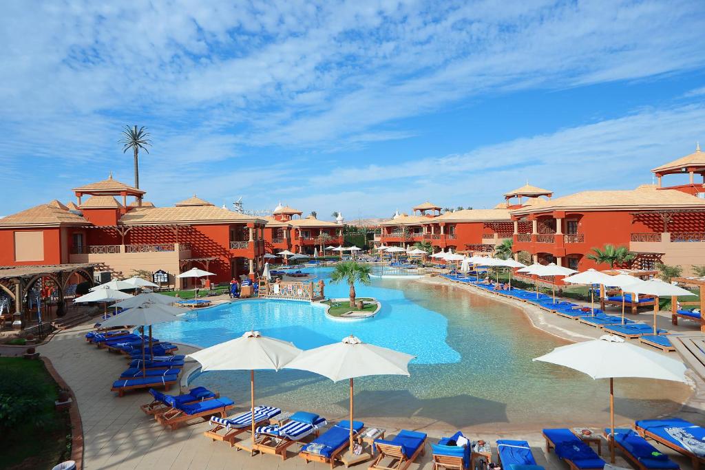 Resort pool area with red buildings, blue loungers, and umbrellas under a clear sky