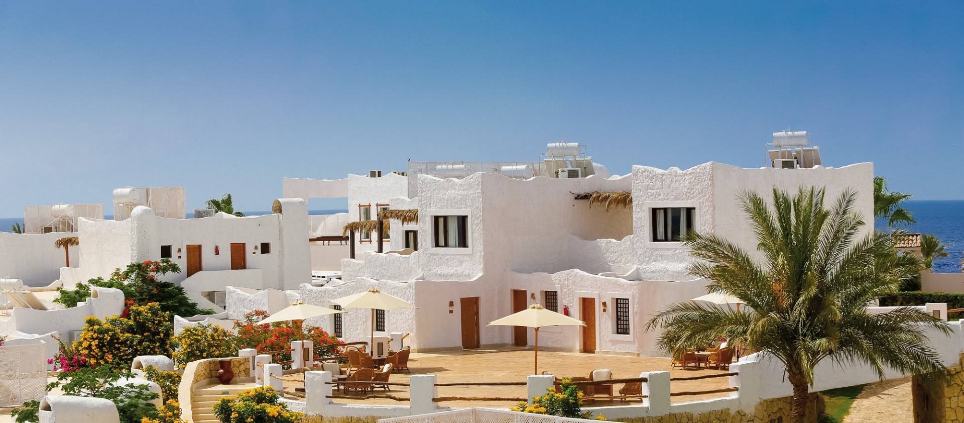 White Mediterranean-style resort buildings with palm trees and colorful flowers under clear blue sky