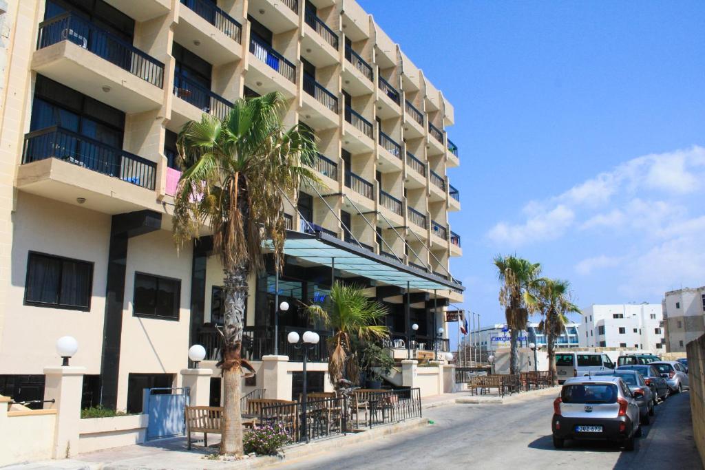 Street view of the hotel exterior with balconies, palm trees, terrace seating, and parked cars.