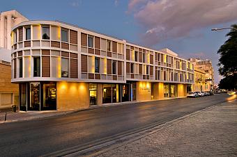 Moderne Hotelaußenansicht an einer Stadtstraße bei Dämmerung mit beleuchteter Fassade und Eingang.