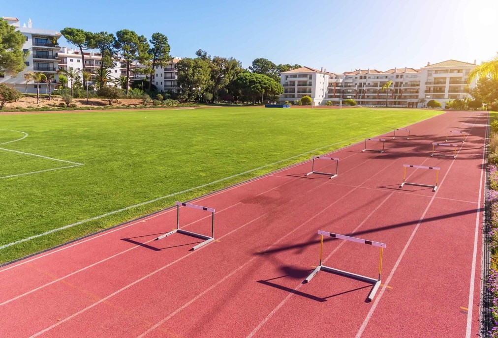 Laufbahn mit aufgebauten Hürden neben Rasenfeld und Hotelgebäuden.