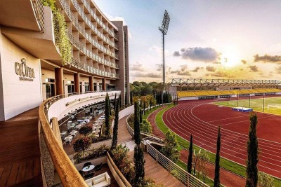 Stimmungsvolle Ansicht des Gloria Sports Arena Gebäudes neben Laufbahn und Tribünen, im warmen Abendlicht. Terrassen und Grünanlagen verbinden das Hotel direkt mit den Sportanlagen.