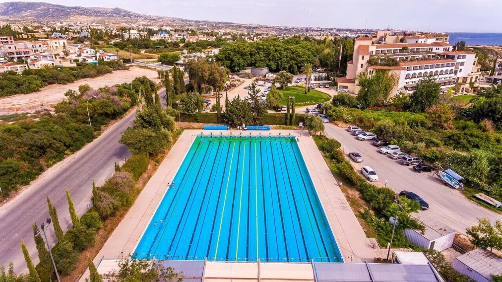 Luftaufnahme eines Langbahn-Schwimmbeckens mit Parkplatz und Landschaft im Hintergrund