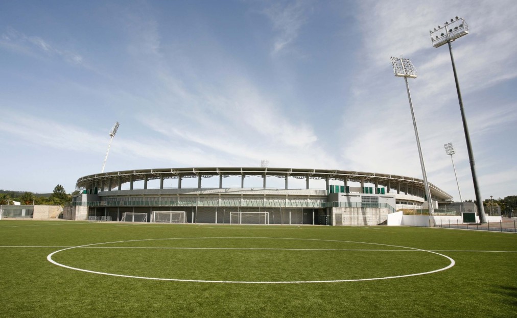Blick über einen Fußballplatz auf ein modernes Stadiongebäude mit hohen Flutlichtmasten.