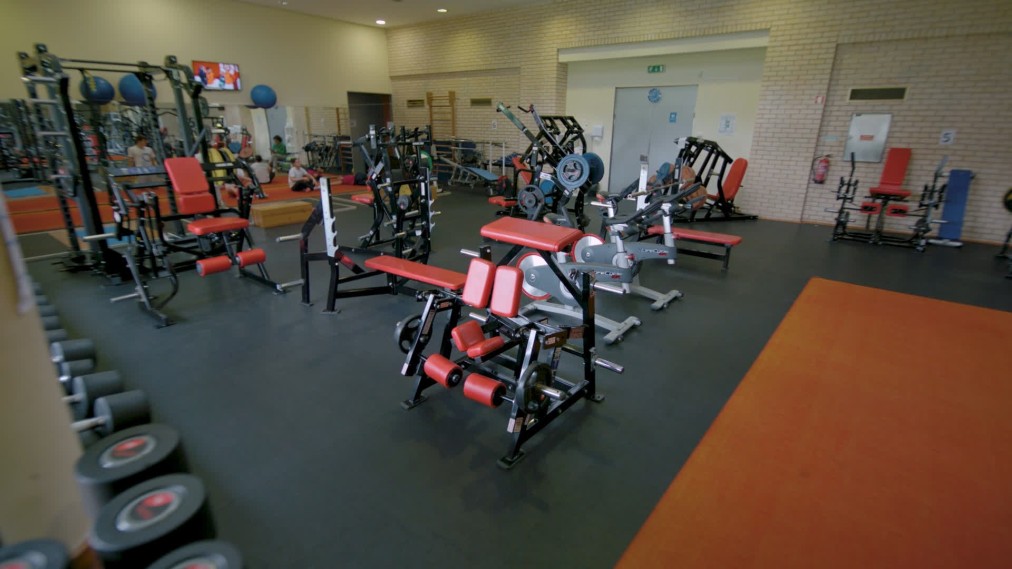 Kraftraum mit Trainingsgeräten, Bänken, Hanteln und Athleten während des Trainings.