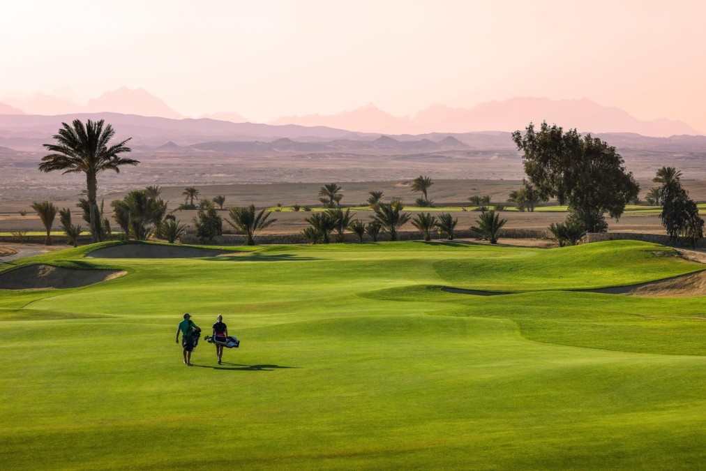 Zwei Golfer gehen über ein Fairway mit Tragetaschen, Palmen und Bergen in der Abenddämmerung.