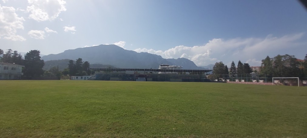 Leeres Fußballrasenfeld mit Toren, Tribüne und Bergen unter klarem Himmel.