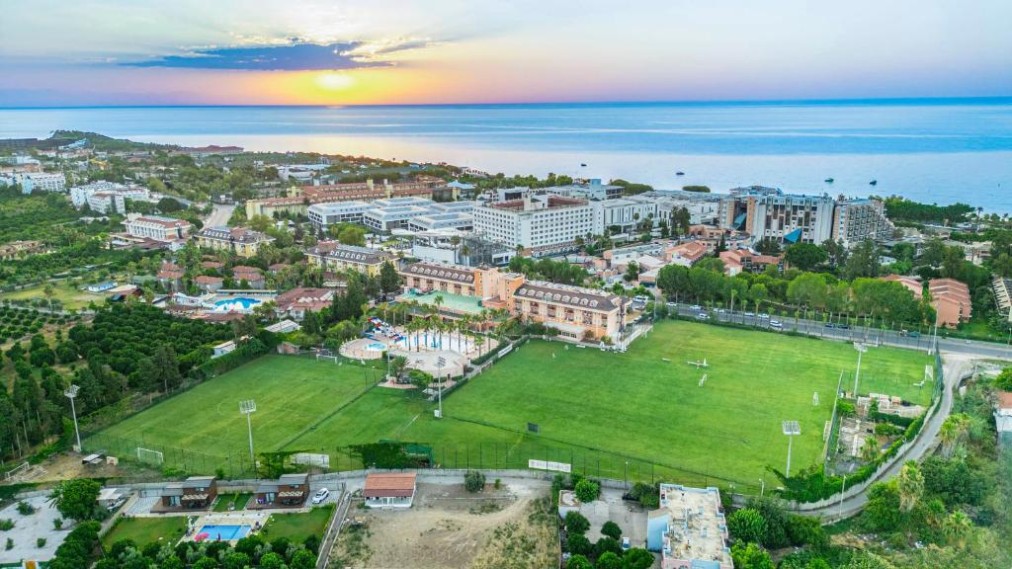 Luftaufnahme bei Sonnenuntergang mit Hotelanlage und Fußballplatz mit Blick aufs Meer.