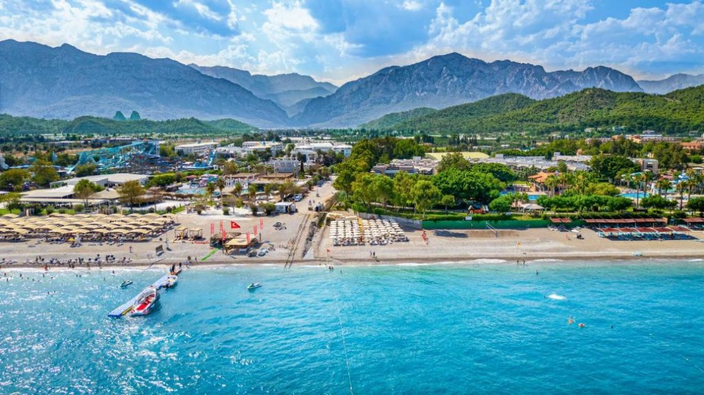Luftaufnahme der Strandpromenade mit Liegen, Steg und Bergen hinter der Hotelanlage.